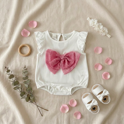 Flat lay arrangement of the white baby romper with pink bow alongside cute summer accessories, baby shoes, and wooden toys on a beige linen backdrop.