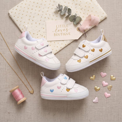 Flatlay display of white baby girl sneakers showing pastel, gold, and multi-colored heart print variants arranged beautifully with decorative props and loose heart confetti.