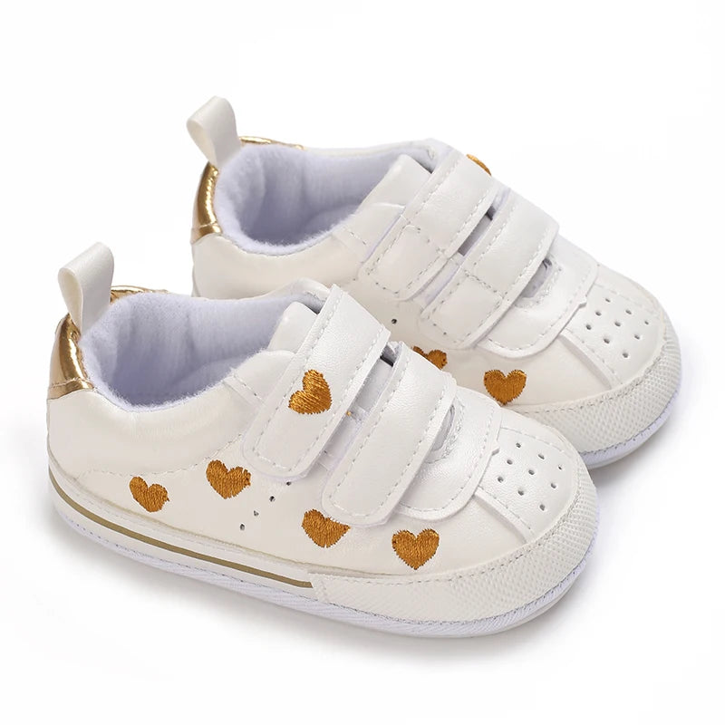 Pair of white baby girl sneakers decorated with gold embroidered hearts, perforated details, and double adjustable straps.