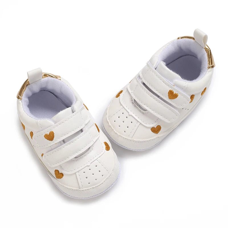 Top-down view of white infant sneakers featuring a metallic gold heart print pattern and gold heel accents.