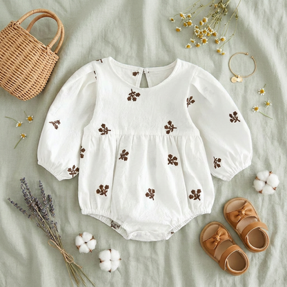 Flat lay of the white floral embroidered long-sleeve baby romper, beautifully styled with brown leather bow sandals, a mini woven basket, cotton bolls, and dried flowers on a soft green linen background.