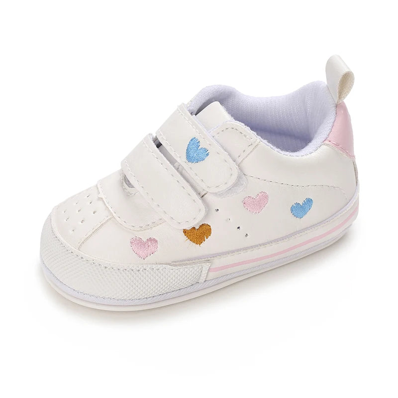 Side profile of a single white baby sneaker displaying pastel embroidered hearts, perforated toe details, and a pink heel tab.