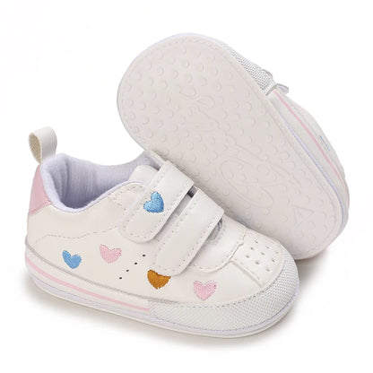 Pair of white baby sneakers with multi-colored hearts, with one resting on its side to display the flexible white anti-slip sole.