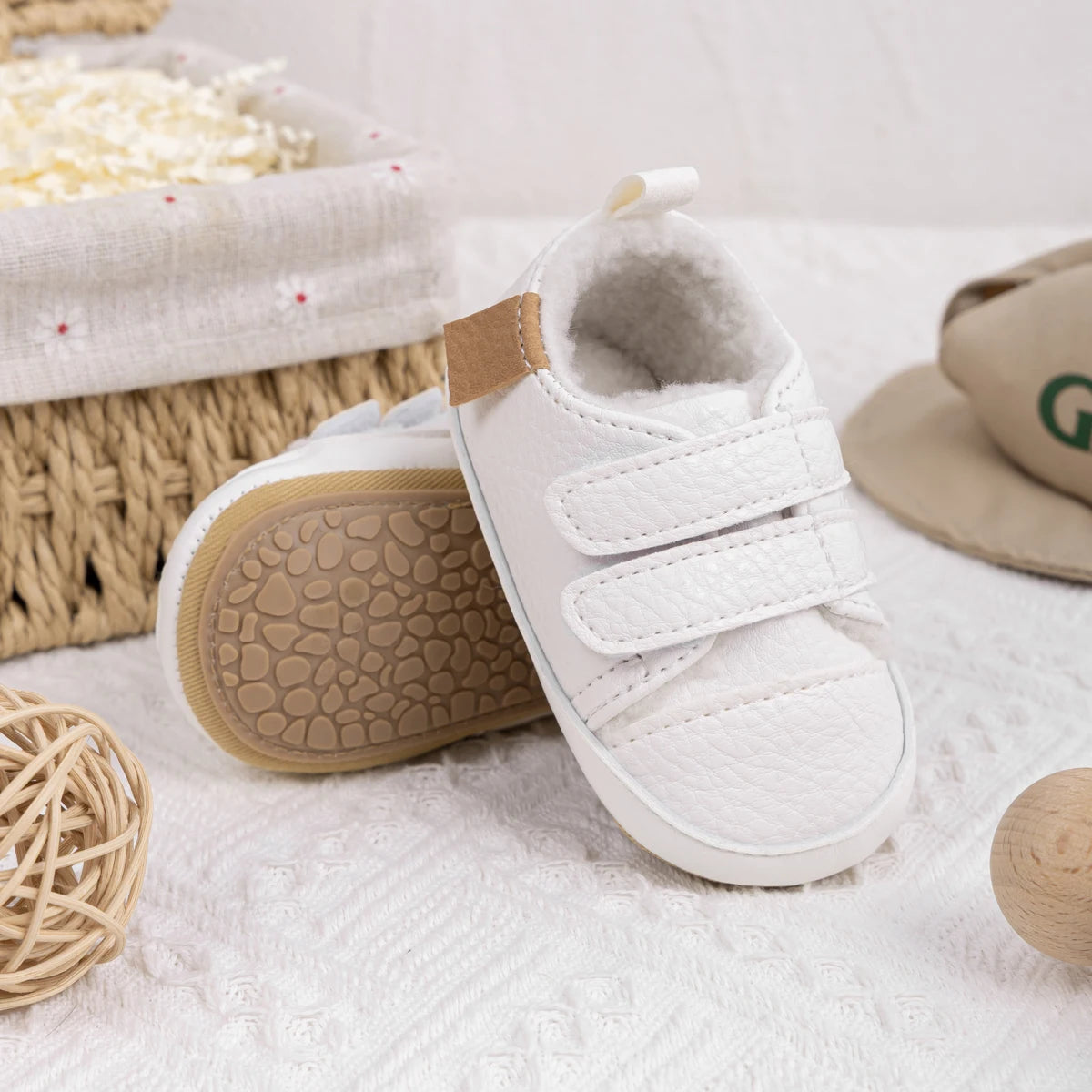 One white fleece-lined infant sneaker standing upright showing double adjustable straps, next to a second shoe lying on its side to display the tan, pebble-textured anti-slip bottom sole.