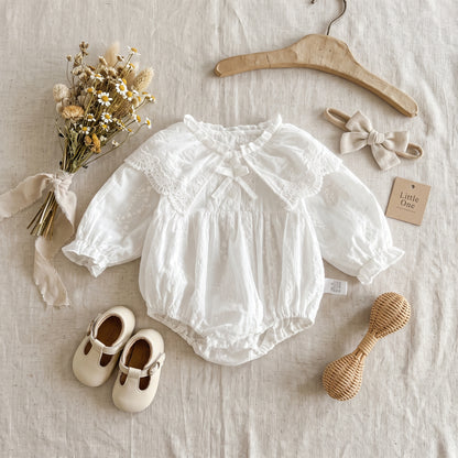 Flat lay of white infant lace bubble romper displayed with a wooden hanger, baby shoes, and dried flowers