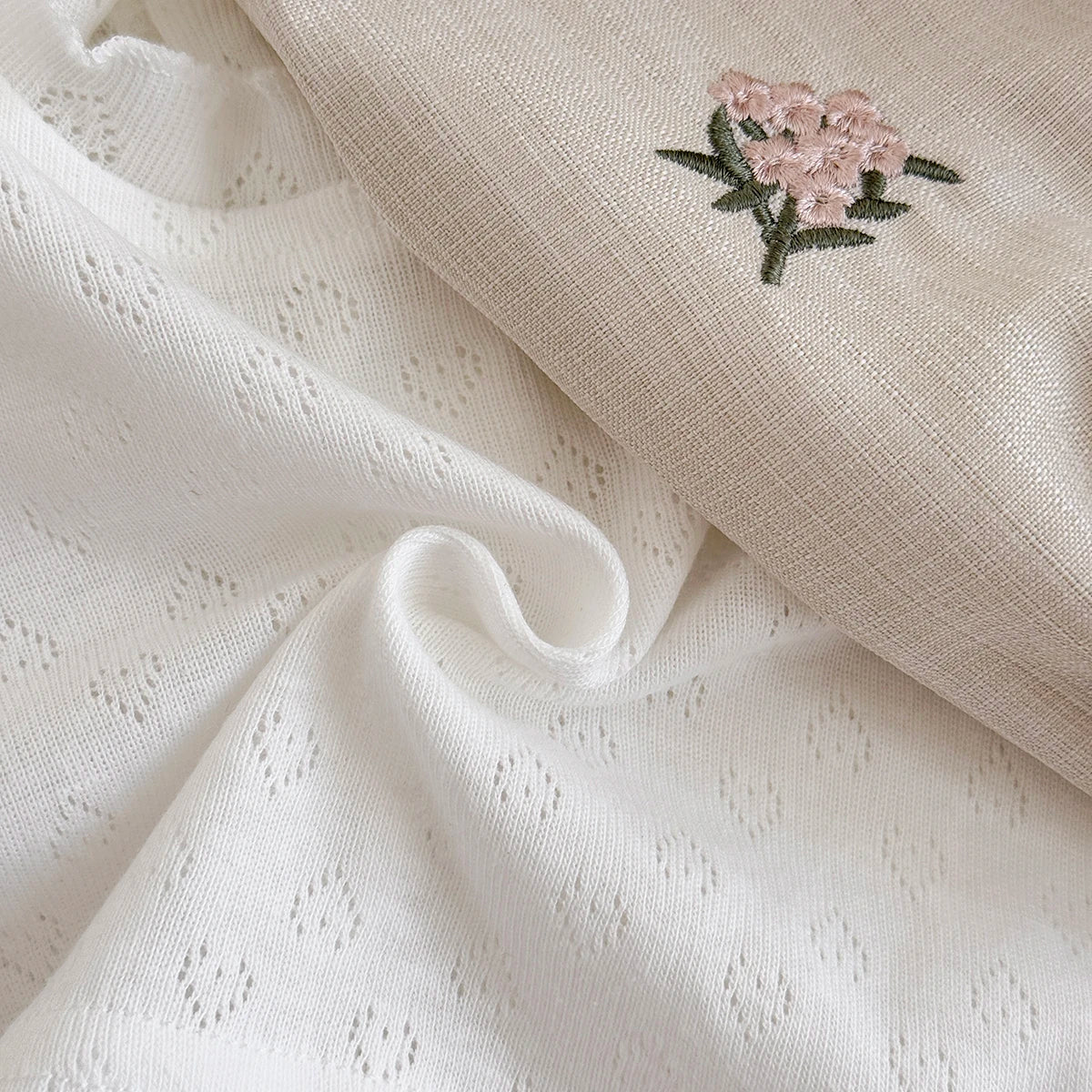 Close-up texture shot showing the soft white heart-shaped pointelle knit fabric next to the woven beige linen-style romper material with a stitched flower.