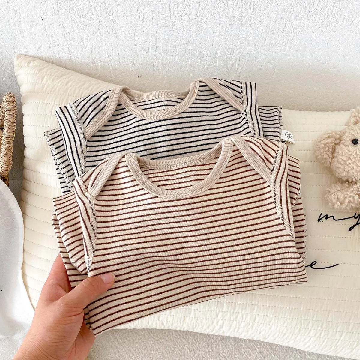 A hand holding neatly folded soft cotton striped infant jumpsuits in both Toasted Latte and Oat & Onyx colors.