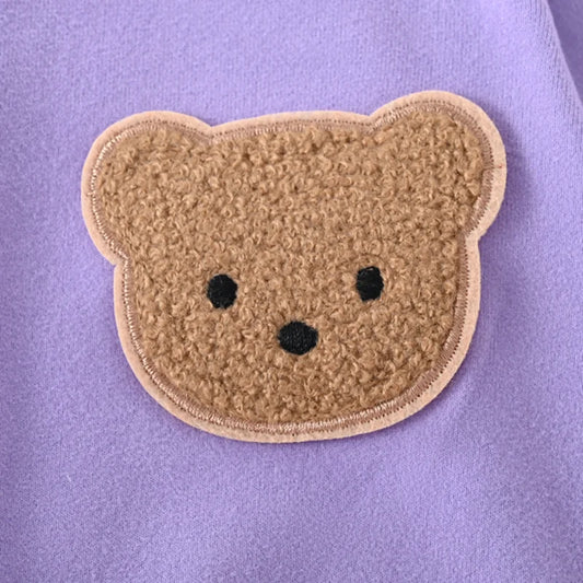 Close-up detail showing the textured brown teddy bear patch applique sewn onto the purple fabric of the newborn kimono romper.