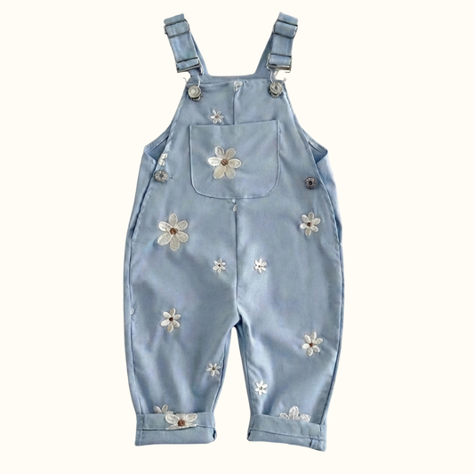 Flat lay of light blue toddler overalls featuring white daisy embroidery, adjustable metal buckle straps, a front bib pocket, and rolled cuffs against a textured white background.