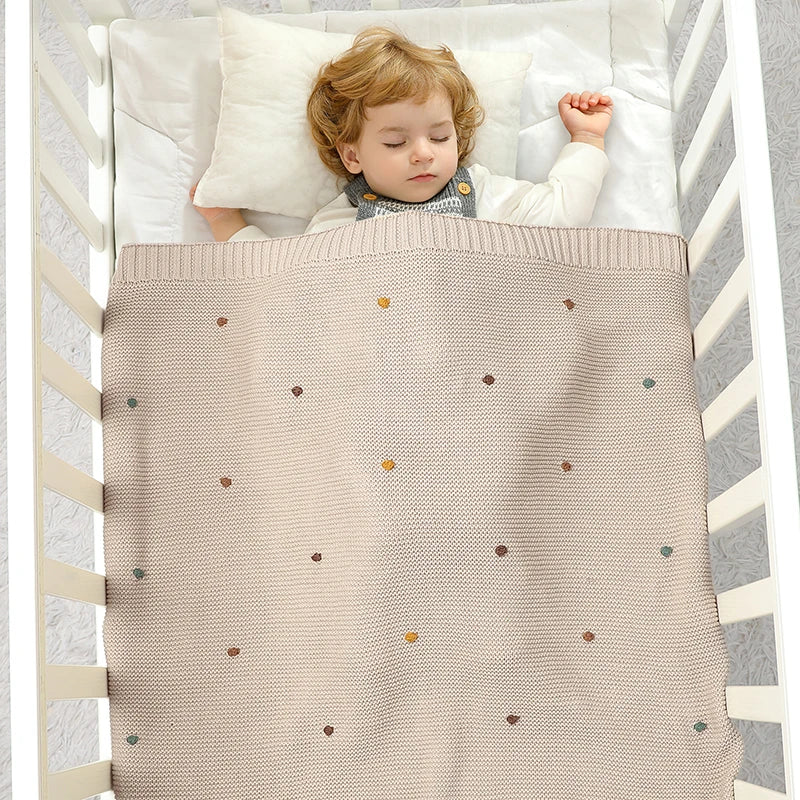 Overhead view of a baby sleeping peacefully in a white wooden crib, covered by the neutral Toasted Almond 100% cotton knitted blanket with multicolored decorative pips.