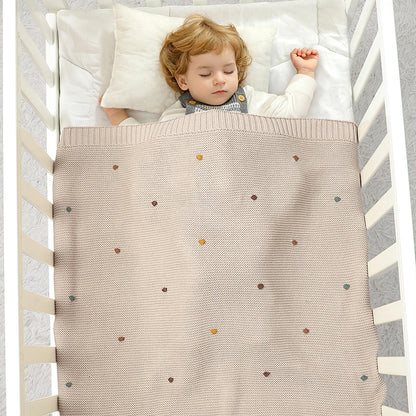 Overhead view of a baby sleeping peacefully in a white wooden crib, covered by the neutral Toasted Almond 100% cotton knitted blanket with multicolored decorative pips.